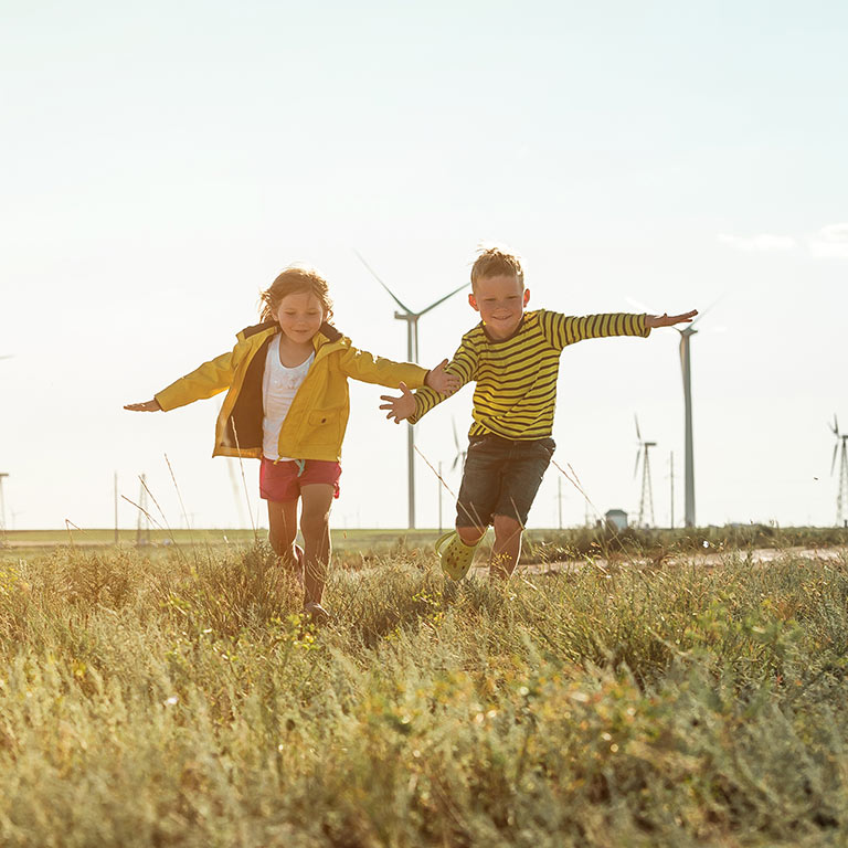 The photo shows two happy children playing in a field. In the background you can see several wind turbines
